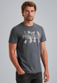 Camiseta de algodón gris con un estampado gráfico que incluye el texto "PME Legend" y otros detalles. Mangas cortas y cuello redondo. Combinada con jeans oscuros.
