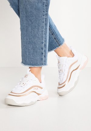 Stopy w białych i różowozłotych butach FILA w połączeniu z niebieskimi jeansami o krótkiej długości na jednolitym jasnym tle.