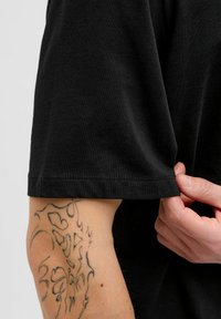 Sort t-shirt med en blød bomulds tekstur, der har en lige kant og en optrullet ærme, der viser en del af armen med et tatoveringsdesign.