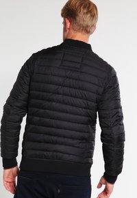 Musta toppatakki, jossa on tikattu pinta, resorit hihansuissa ja helmassa, kapean mallinen muotoilu ja ei näkyvää bränditystä.