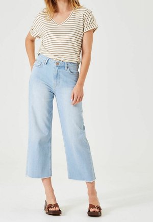 Frau, die ein beige-weiß gestreiftes, kurzärmliges Oberteil, hellblaue, weit geschnittene, gekürzte Jeans und braune Sandalen mit Absatz trägt und mit einer Hand in der Hüfte steht.