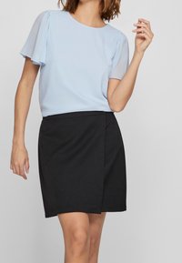 Blusa azzurra chiara con maniche corte e trasparenti abbinata a una gonna nera a campana, caratterizzata da una texture liscia e un design semplice.