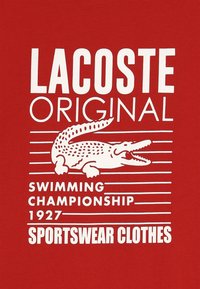 T-shirt rossa con testo bianco che recita "Lacoste Original," accompagnato da un grafico di un coccodrillo e strisce orizzontali. Il testo include "Campionato di Nuoto 1927."