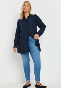 Camicia blu navy con bottoni sul davanti, maniche lunghe e motivo ad occhiello, abbinata a jeans skinny blu chiaro e ballerine nere.