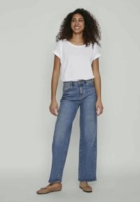 Woman with curly hair wearing a white short-sleeve shirt, high-waisted blue jeans, and beige flats, standing and smiling against a plain background.