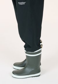 Bottes de pluie en caoutchouc vertes avec une bande blanche près du haut, semelle texturée, associées à un pantalon imperméable noir.
