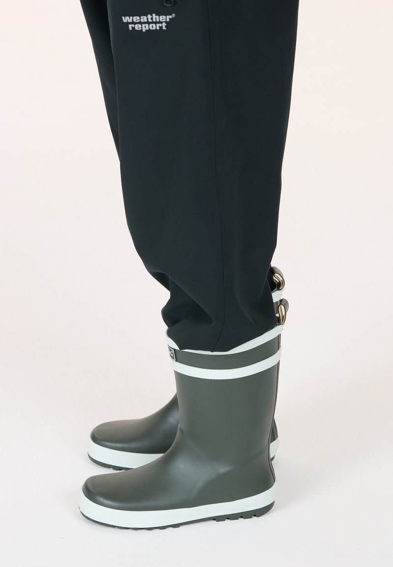 Bottes de pluie en caoutchouc vertes avec une bande blanche près du haut, semelle texturée, associées à un pantalon imperméable noir.
