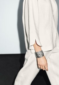 Bracelet en argent avec un design texturé porté sur une main, associé à un haut en lin léger avec des manches fendues et une coupe décontractée.