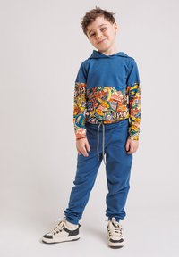 Młody chłopiec uśmiecha się, ubrany w niebieską bluzę z kapturem ozdobioną kolorowymi naszywkami sportowymi, dopasowane niebieskie spodnie oraz białe tenisówki z czarnymi akcentami.