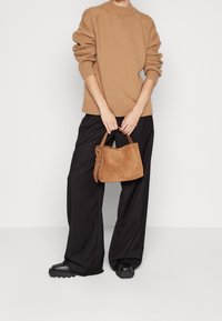 Pull en maille marron avec un col côtelé, associé à un pantalon noir à jambes larges. Tenant un petit sac à main en daim marron avec une finition texturée.