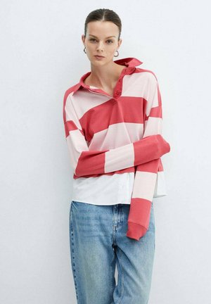 Tricou polo cu mânecă lungă, cu dungi roșii și roz, cu guler, purtat peste o cămașă albă, asortat cu blugi de culoare albastru deschis.