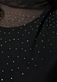 Tessuto nero con un sovrapposto trasparente, decorato con piccoli strass disposti in modo casuale, creando un effetto scintillante sottile sulla superficie.