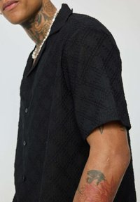 Chemise noire à manches courtes présentant un motif en diamant texturé, un design à col et des boutons sur le devant, offrant une coupe décontractée.