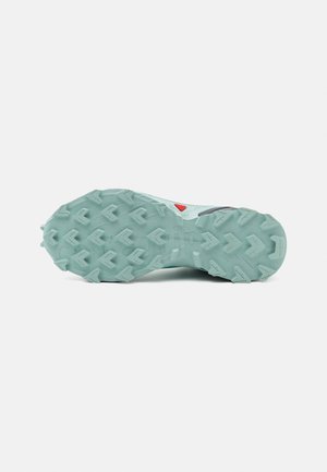 Semelle d'une chaussure de trail running vert clair avec des crampons multidirectionnels et un petit détail triangulaire rouge près de la voûte plantaire.