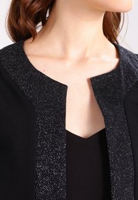 Czarna kurtka z błyszczącą czarną lamówką. Dekolt jest odsłonięty, prezentując warstwowe tkaniny o gładkiej fakturze i błyszczących detalach na krawędziach.