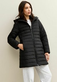 Piumino nero con design a righe orizzontali, cappuccio, maniche lunghe e chiusura fronte con zip. Tessuto leggero con una texture liscia.