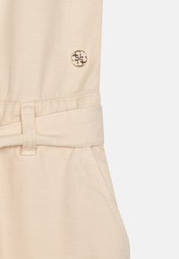 Tissu en mélange de coton beige présentant une taille ceinturée, des coutures diagonales et un détail logo métallique circulaire sur le côté.