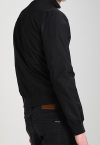 Svart bomberjacka med hög krage, elastiska cuffs och nederkant. Slät textur, minimalistisk design, har en etikett på baksidan.