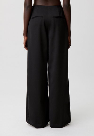 Pantalones negros de pierna ancha con tejido suave, cintura alta y dos bolsillos traseros con costuras sutiles. Corte entallado, largo hasta el suelo.