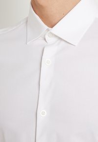 Acercamiento de una persona que lleva puesta una camisa blanca de vestir, bien abotonada, con cuello puntiagudo.