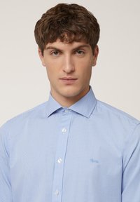 Light blue checkered shirt with a button-down collar and white buttons. Features a small embroidered logo on the left chest. Smooth texture.