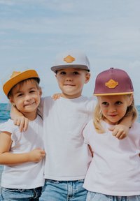 Three children wearing baseball caps: gray with orange brim, purple with yellow brim, and tan patches; light shirts and denim shorts.
