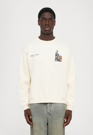 Young man wearing off-white sweatshirt with stacked vintage cassette tapes graphic and text "Filling Pieces" on chest, paired with faded jeans.