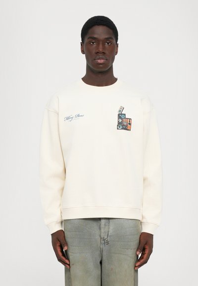 Junger Mann in einem cremefarbenen Sweatshirt mit einem gestapelten Grafik-Motiv von Vintage-Cassetten und dem Text "Filling Pieces" auf der Brust, kombiniert mit ausgewaschenen Jeans.