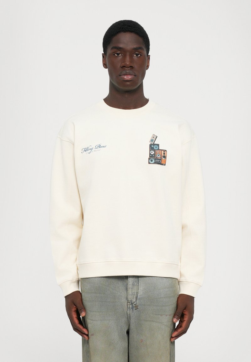 Jovem vestido com um sweatshirt bege claro com uma gráfica de cassetes vintage empilhados e o texto "Filling Pieces" no peito, combinado com jeans desbotados.