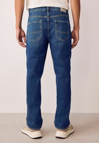Jeans din denim cu picioare drepte, spălare medie și buzunare pe spate. Prezintă cusături în contrast și un talie standard, purtate cu adidași deschisi la culoare.