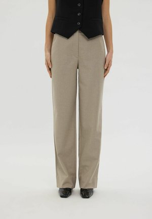 Personne portant un pantalon beige à jambes larges, des chaussures noires pointues et un gilet noir boutonné, debout devant un fond clair uni.
