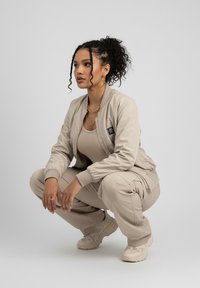 Beige Bomberjacke mit gerippten Bündchen, dazu passende lässige Cargo-Hosen und neutrale Sneaker. Merkmale sind Reißverschlussdetails und geschichteter Schmuck.