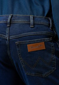Dark blue denim jeans with a brown leather Wrangler label on the back pocket, featuring classic stitching and a tapered fit design.