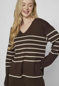 Pull long à rayures marron avec un décolleté en V. Il présente des rayures horizontales crème et une coupe décontractée, fabriqué en matière tricotée douce.