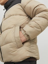 Giacca a vento beige con parte superiore nera, dotata di chiusura con zip, polsini elasticizzati e tasche laterali. Texture liscia e trapuntata.
