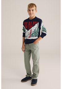 Jūras zila adīta džemperis ar sarkanu un baltu kalnu grafikā, zaļiem kokiem un snovotāja detaļām. Saskaņots ar gaiši zaļām biksēm un jūras zila apaviem.