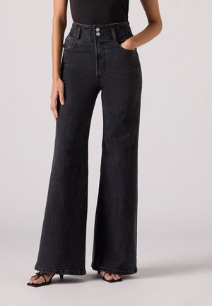 Vrouw staand, gekleed in zwarte high-waist wijde jeans en zwarte sandalen met hak en bandjes, één hand in de zak, tegen een effen achtergrond.