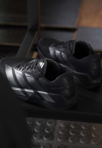 Μαύρα παπούτσια τρεξίματος Adidas Adizero DropSet με αντανακλαστικές ρίγες τοποθετημένα σε ένα σκοτεινό παγκάκι σε χαμηλά φωτισμένο εσωτερικό χώρο.