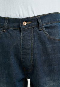 Los vaqueros de mezclilla azul oscuro cuentan con un diseño clásico de cinco bolsillos, con costuras naranjas en contraste y un cierre de botón de cobre en la cintura.