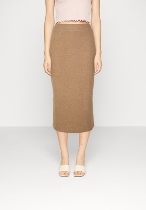 Vrouw draagt een beige geribbelde midi kokerrok, een lichtroze cropped top en witte openhakken, en staat voor een effen achtergrond.