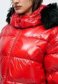 Piros, fényes felületű pufi dzseki, magas gallérral és fekete szőrmével díszített. Mintás steppeléssel és elöl cipzáras részletekkel.