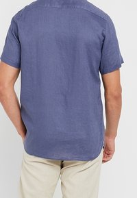 Camisa de lino de manga corta, azul oscuro, con cuello y yugo en la parte posterior; presenta un dobladillo recto y aberturas laterales. Combinada con pantalones beige claros.