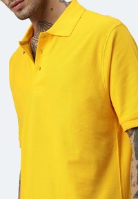 Gelbes Polo-Shirt aus strukturiertem Baumwolle, mit klassischem Kragen und Knopfleiste mit zwei Knöpfen, schräg präsentiert.