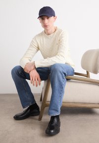 Cream cable-knit sweater, blue jeans, black slip-on shoes, and a navy cap. Seated on a light upholstered chair with wooden legs.