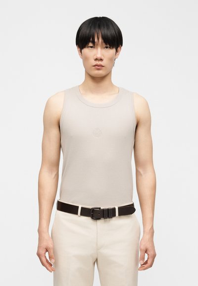 Άνδρας με ίσια μαύρα μαλλιά, ντυμένος με μπεζ αμάνικο τοπ και ανοιχτόχρωμο παντελόνι, στέκεται μπροστά σε απλό ανοιχτό φόντο.