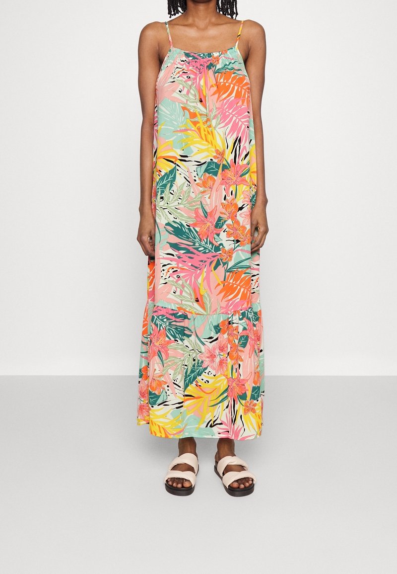 Femme portant une robe longue sans manches à imprimé tropical coloré avec fines bretelles et sandales blanches à enfiler, debout devant un fond uni.