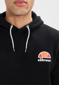 Hombre vistiendo una sudadera negra con cordones blancos y un logo de ellesse en naranja-rojo en el pecho, fondo claro neutro.