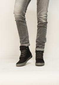 Botas altas de ante negras con suelas de goma gris, con cierre de cordones y un sutil detalle de costura, combinadas con unos jeans ajustados grises.