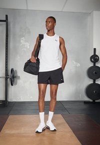 Witte mouwloze tanktop, zwarte shorts met logo, witte sneakers met accenten, een zwarte tas dragend; sportschoolomgeving met gewichten op de achtergrond.