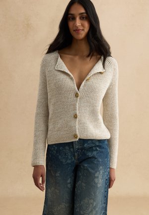 Donna che indossa un cardigan beige lavorato a maglia con bottoni e jeans blu a fantasia floreale, in piedi davanti a uno sfondo beige uniforme.